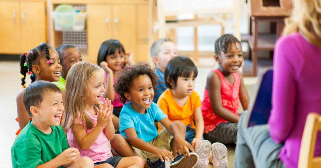 A group of diverse children wearing bright colored t-shirts, listen to an adult read a story