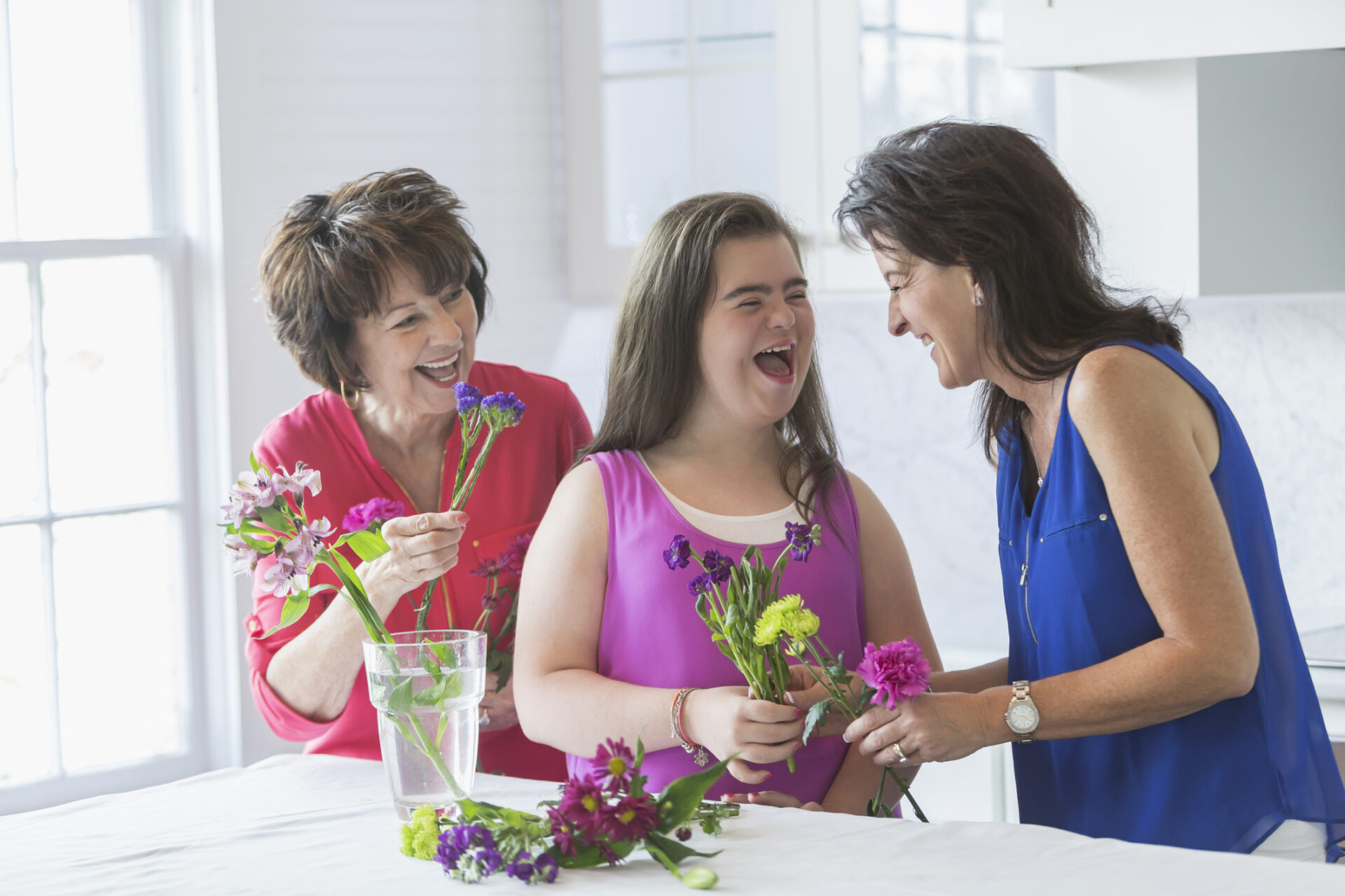 A teenage girl with downs syndrome arranging flowers with her mother and grandmother. They are holding cut flowers in their hands, talking, laughing and looking at mom. They are at home in the kitchen by a bright, sunny window.