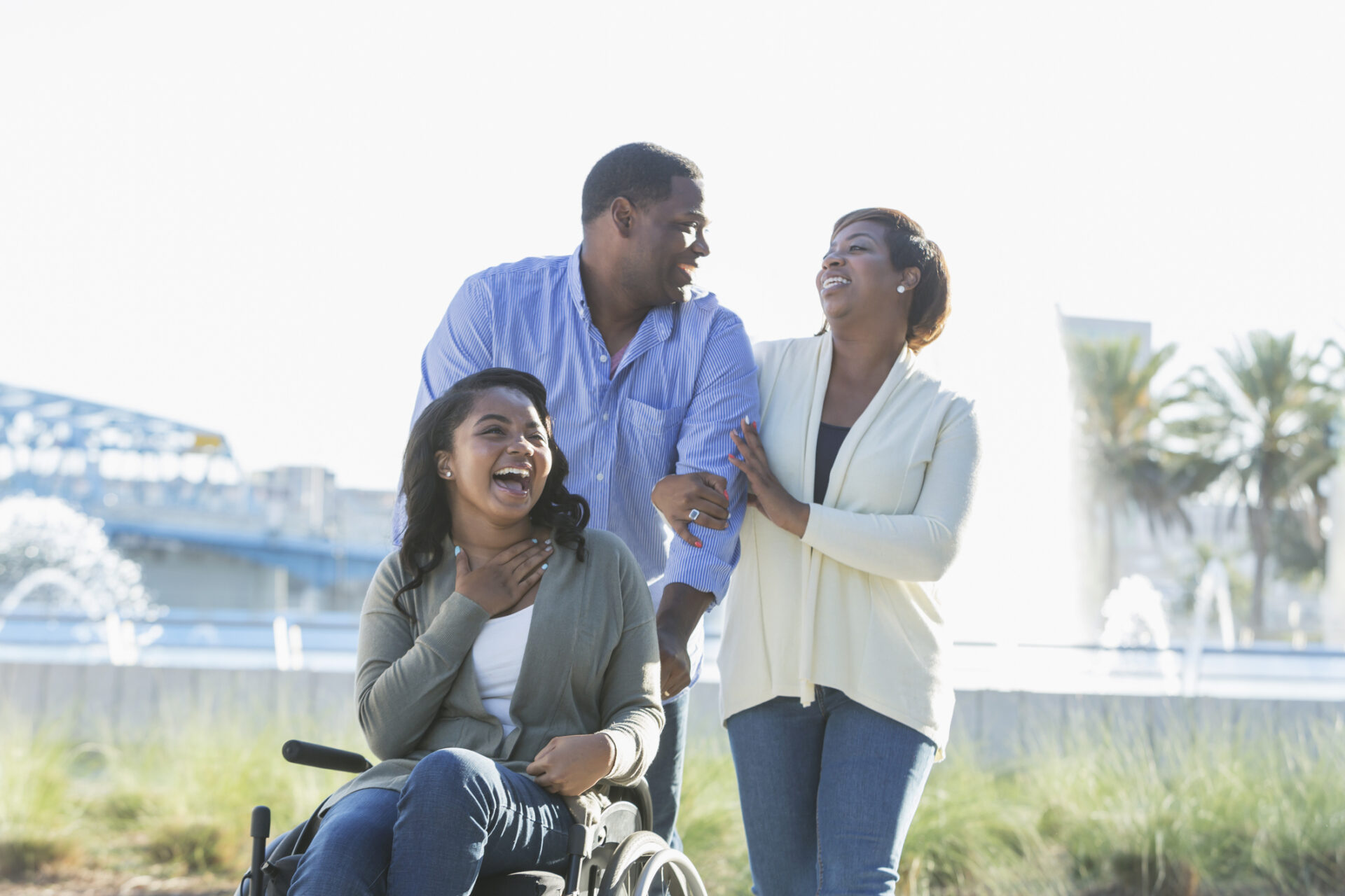 An African American family walking near a city waterfront, buildings and a bridge in the background. The teenage daughter, 15 years old, is a paraplegic, sitting in a wheelchair.